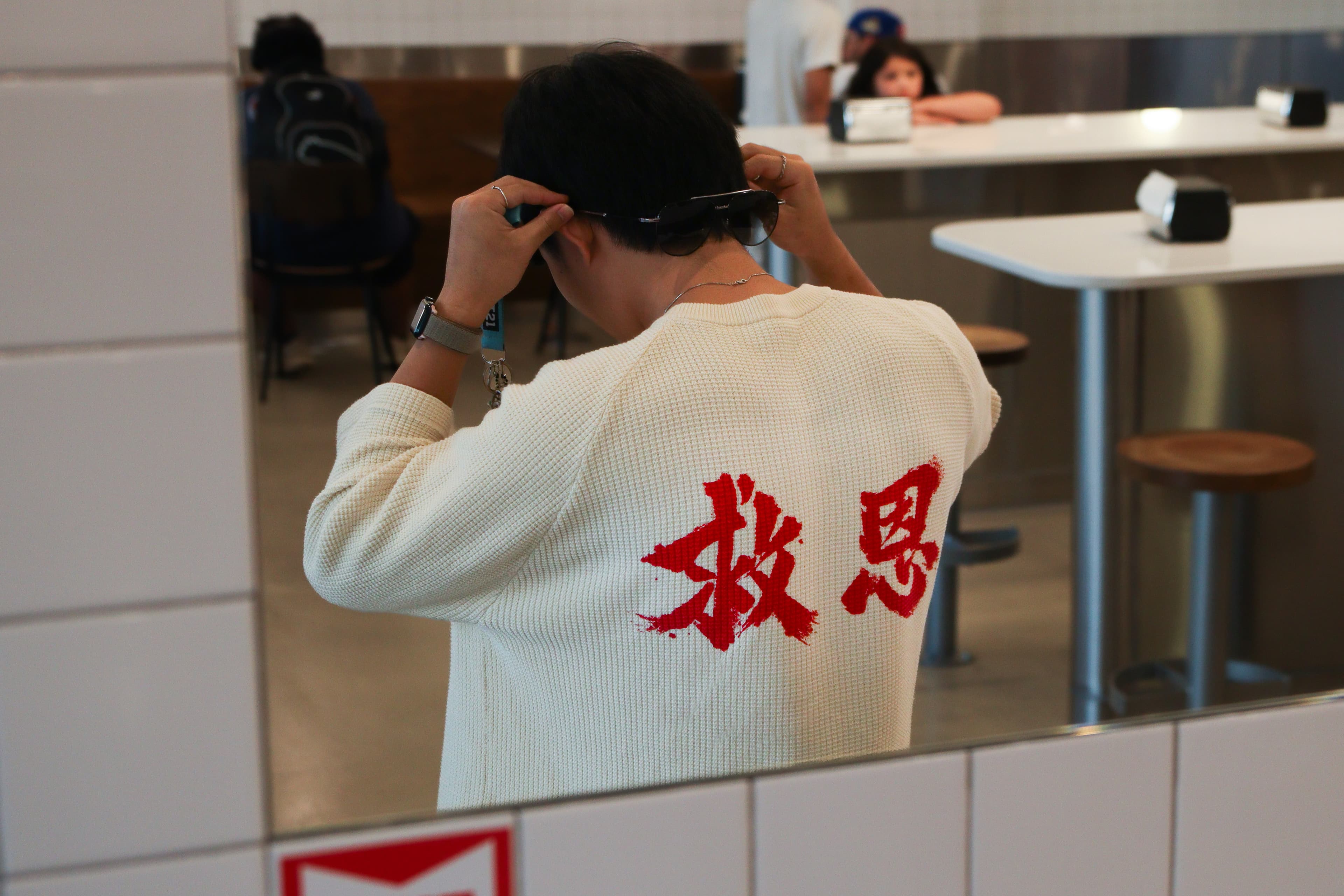 Photographed from behind in a mirror, wearing a cream cardigan with 救恩 (salvation) embroidered across the back in red, adjusting a pair of sunglasses.