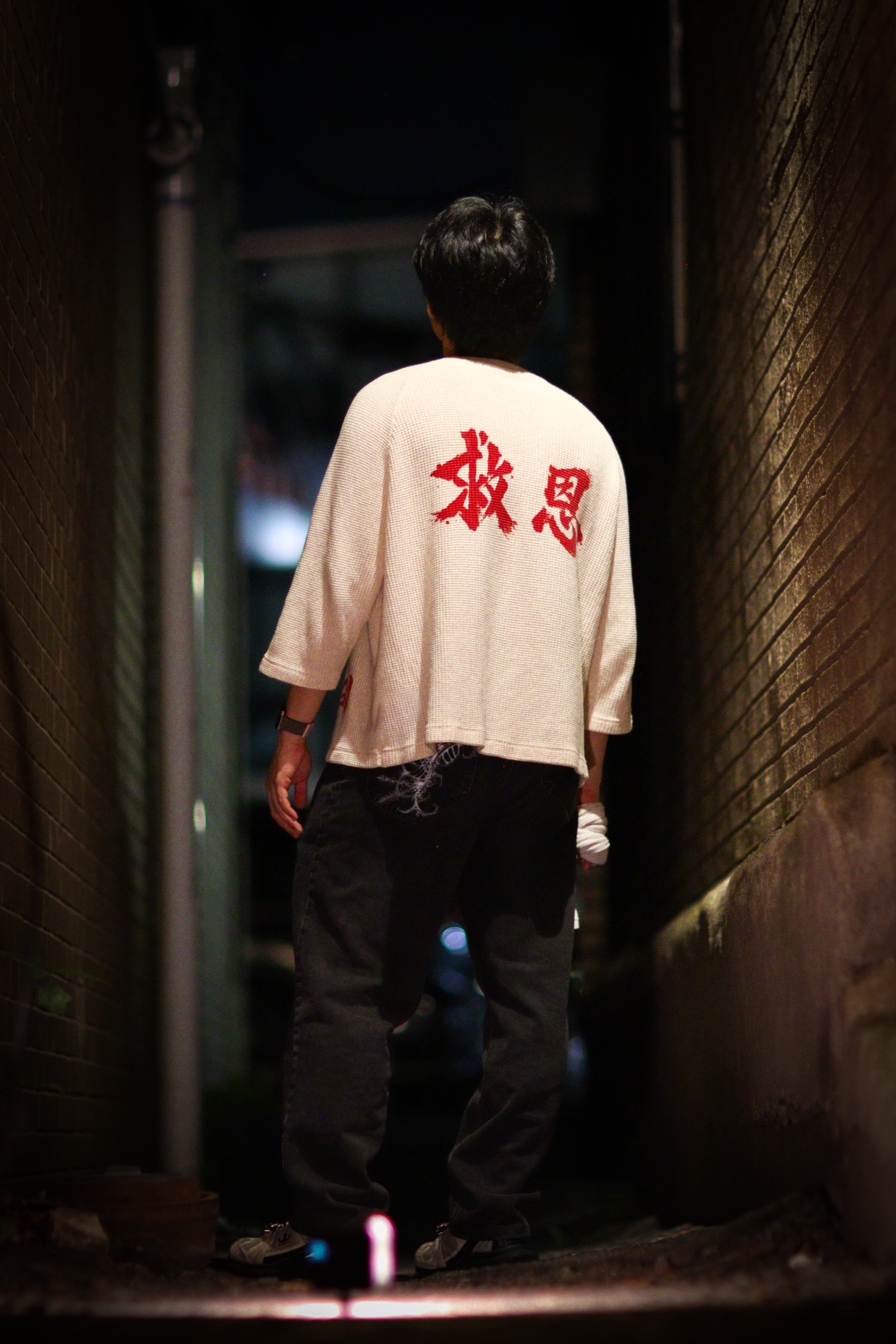 On-model, back, walking away down a narrow alley at night, lit from behind — the 救恩 mark visible on the waffle knit.