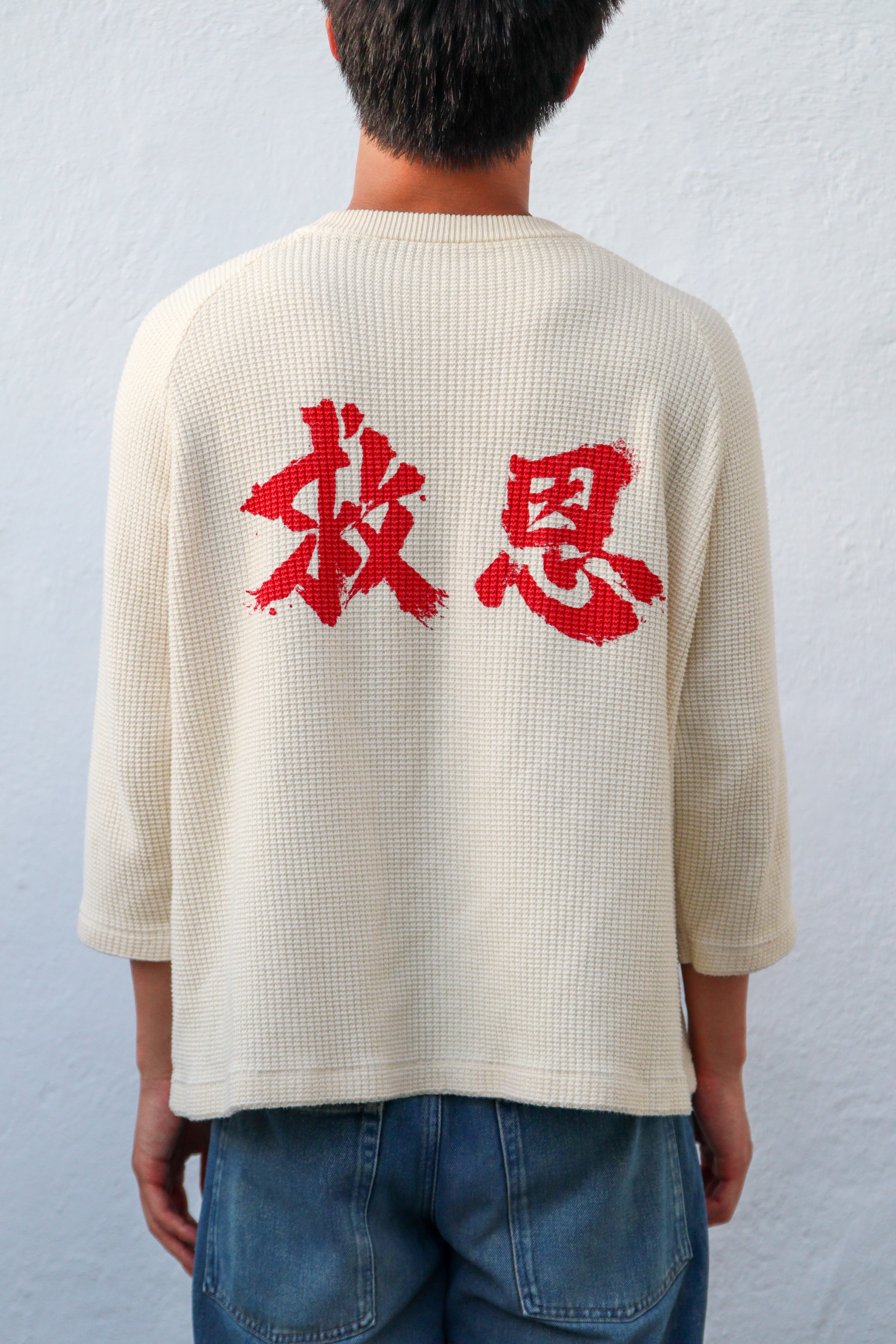 On-model, male, back, straight on, plain studio background: the 救恩 mark in red across the cream waffle knit.